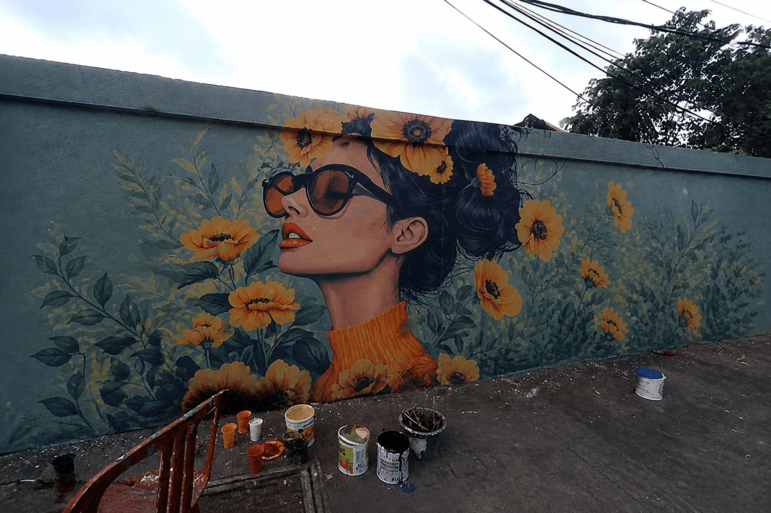 Mural wanita berkacamata dengan bunga warna kuning di tembok luar hasil lukisan tangan